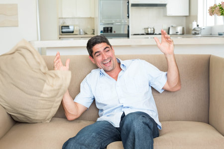 Middle-aged man wearing blue shirt and jeans tossing pillow on beige sofa near kitchen cabinets. Contemporary, casual, cozy, bright, inviting, minimalist, lifestyleの写真素材