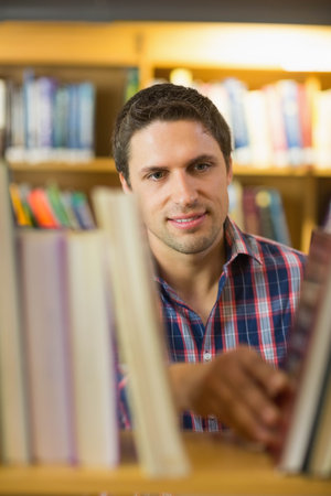 Man wearing plaid shirt browsing colorful books at wooden library bookshelf under warm lighting. Literature, education, reading, academic, interior, knowledge, explorationの写真素材