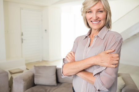 Mature woman standing arms crossed smiling at camera in living room with sofa cushions, copy space. Contemporary, cozy, inviting, elegant, lifestyle, relaxation, interiorの写真素材