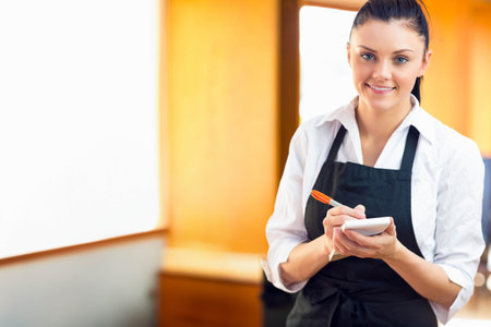 Woman holding pen over order pad while wearing black apron taking order in restaurant, copy space....