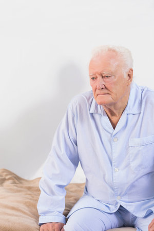 Senior man leaning forward on bed edge in pajamas near beige blanket and mattress, copy space. Senior, retirement, relaxation, comfort, wellness, contemplation, cozyの写真素材