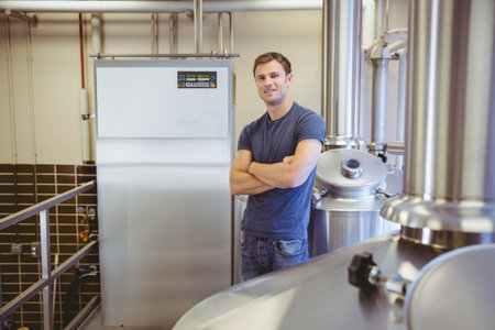Brewer standing with arms crossed beside control panel with digital readout in brewery tanks, pipes. Industrial, craftsmanship, machinery, innovation, precision, productivity, engineeringの写真素材