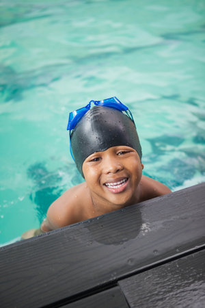 African American child boy leaning on pool deck at turquoise pool wearing black cap, blue goggles. Children, leisure, recreation, aquatic, summertime, playful, vibrantの写真素材