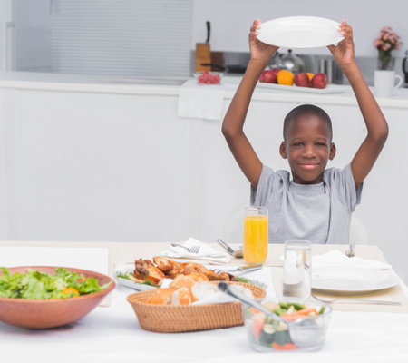 African American boy smiling and holding empty white plate above head at home kitchen table. Joy, youth, nourishment, celebration, cozy, wholesome, freshの写真素材