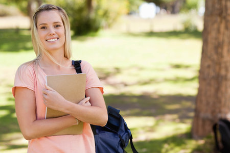 Female student standing on grassy campus quad holding notebook against chest and carrying backpack. Outdoor, education, nature, productivity, youthful, serenity, leisureの写真素材