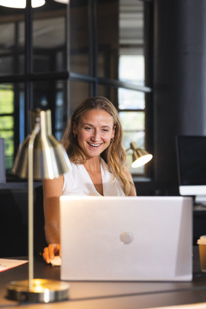 Smiling woman working on laptop at office desk with modern lamp. Business, technology, workplace,...