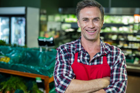 Standing in produce section, male wearing red apron over plaid shirt by wooden bins, copy space. Professional, retail, vibrant, health, freshness, hospitality, visualの写真素材