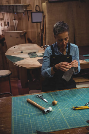 Senior woman wearing apron stitching leather by hand at workshop bench with cutting mat, copy space....