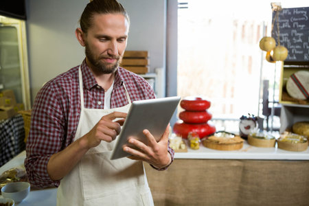 Man using tablet computer behind counter in cheese shop with red-waxed cheese wheels and baskets. Gourmet, artisanal, rustic, food, retail, modern, farm freshの写真素材