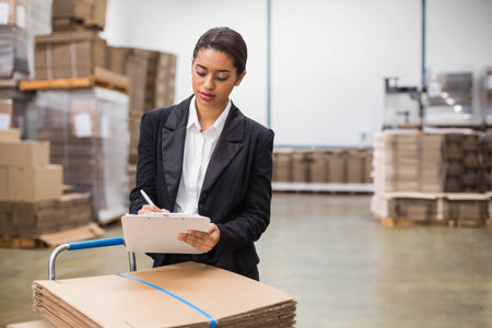 Woman leaning on trolley with cardboard cartons and writing on clipboard near pallets in warehouse. Industrial, logistics, inventory, organizational, modern, distribution, storageの写真素材