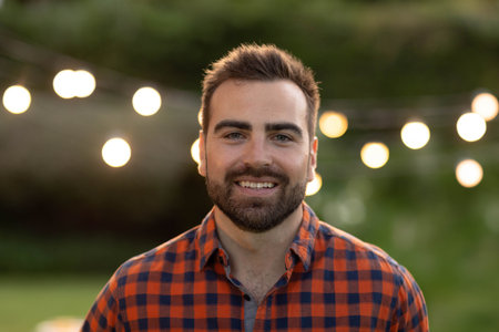 Man standing in backyard garden at dusk wearing checkered shirt under glowing string lights....