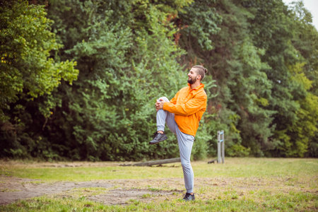 Bearded man in windbreaker performing knee-to-chest stretch in park with exercise post, copy space....