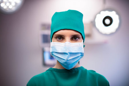 Female surgeon standing in operating room, wearing teal scrubs and mask under glowing lights....