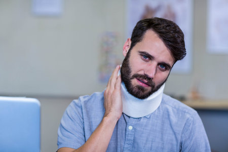 Man sitting at desk at clinic wearing foam neck brace touching neck beside laptop charts. Medical, health, injury, rehabilitation, professional, exam, consultationの写真素材