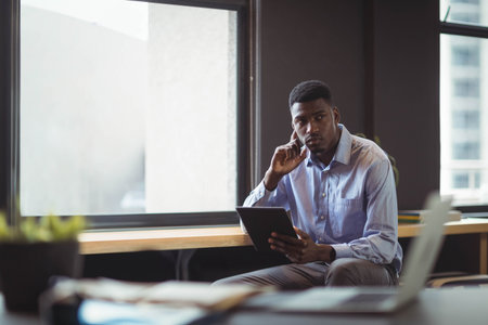 African American man sitting on bench by window in office holding tablet near laptop. Professional,...