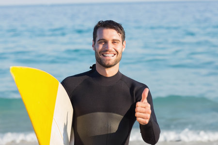 Man smiling and giving thumbs-up standing in sea holding yellow surfboard wearing wetsuit. Adventure, leisure, fitness, outdoor, wellness, sport, vibrantの写真素材