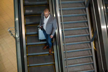 Man standing on down escalator with glass rail at terminal holding suitcase in blazer, copy space. Business traveler, urban, modern, professional, transportation, sleek, movementの写真素材