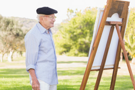 Senior man painter studying blank canvas on easel in grassy park wearing beret, glasses, copy space. Artistic, leisure, serenity, outdoors, landscape, creativity, inspirationの写真素材