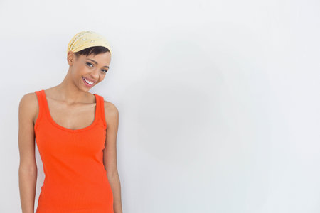 African American woman standing in orange tank top, yellow bandana against white wall, copy space. Portrait, diversity, casual, vibrant, optimistic, femininity, minimalismの写真素材
