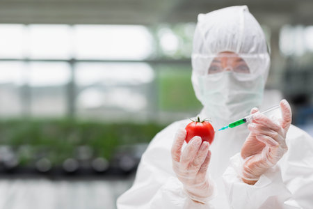 Scientist wearing protective suit injecting red tomato with green liquid in greenhouse, copy space. Research, biotechnology, innovation, laboratory, greenhouse, experimentation, scientificの写真素材