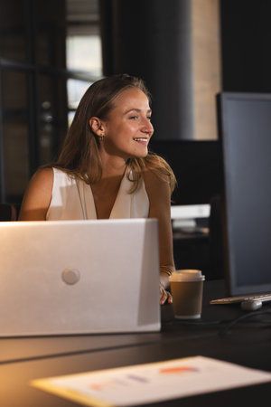 Smiling woman working late at office with laptop and coffee, feeling motivated. Overtime, dedication, focus, productivity, caffeine, technologyの写真素材