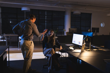 Colleagues discussing project in office at night, analyzing data on computer screen. Collaboration,...