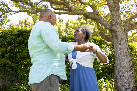Senior couple dancing joyfully together in sunny garden under tree. Elderly, happiness, outdoors, retirementの写真素材
