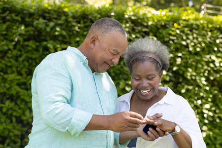 Exchanging rings and smiling, African American senior couple celebrating love in garden. celebration, relationship, happiness, romance, elderlyの写真素材