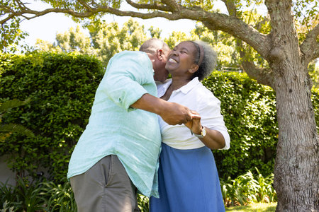 African American senior couple dancing joyfully under tree in sunny garden. Elderly, bonding, relationship, nature, outdoors, leisureの写真素材