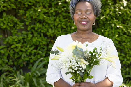 Senior woman holding bouquet of flowers, smiling peacefully in garden. Gardening, nature, floristry, leisure, happiness, outdoorsの写真素材