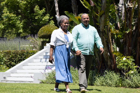 Senior couple walking hand in hand through lush garden, enjoying sunny day. Gardening, nature, outdoors, togetherness, happiness, leisureの写真素材