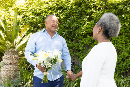 African American man giving flowers to woman in garden, both smiling warmly. Romance, couple, happiness, affection, outdoors, natureの写真素材