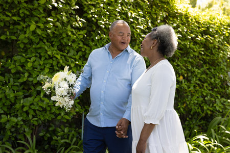 Senior couple holding hands and smiling while walking in garden with flowers. Elderly, gardening, romance, nature, outdoors, happinessの写真素材