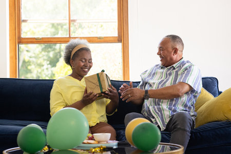 Senior couple exchanging gifts on couch, smiling and enjoying celebration at home. family,...