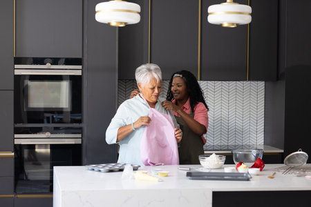 Granddaughter helping grandmother put on apron in modern kitchen, preparing to bake. Family, bonding, baking, assistance, preparation, loveの写真素材