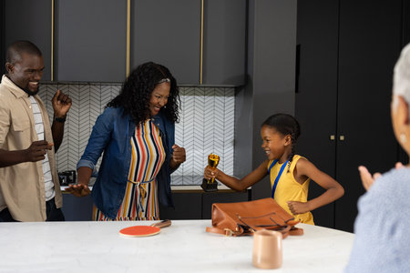 Celebrating at home, family cheering as young girl holds trophy with joy. Celebration, achievement, victory, happiness, support, successの写真素材