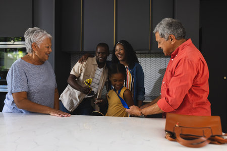 Three generations celebrating young girl's achievement with trophy in modern kitchen. Celebration, happiness, family, success, multigenerational, indoorsの写真素材