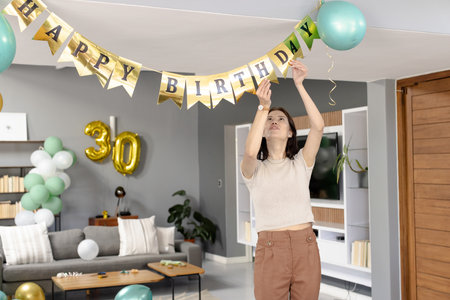 Woman hanging HAPPY BIRTHDAY banner in living room with gold 30 balloon and mint-green balloons. Modern, minimalist, residential, sofa, shelving, coffee table, decoratingの写真素材