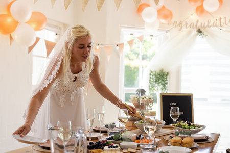 Bride wearing lace gown and veil arranging dishes on table in indoor reception with peach balloons. Wedding, bridal, romantic, festive, party, buffet, cateringの写真素材