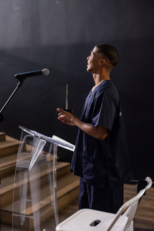 Man standing behind podium with microphone on stage holding trophy smiling upward, copy space. Award, recognition, achievement, celebration, speaker, lectern, corporateの写真素材