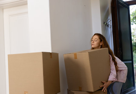 Woman carrying large cardboard box in home entryway with coat hooks and moving boxes, copy space. Relocation, domestic, real estate, new beginning, unpacking, threshold, cartonsの写真素材
