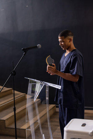 Man standing on stage at clear podium with microphone, holding gold-black award trophy, copy space....