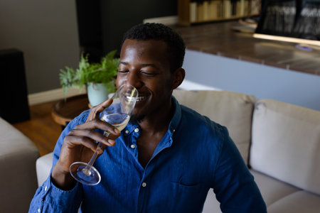 African American man drinking white wine from glass on beige couch in modern living room. Sipping, relaxation, minimalist, contemporary, sofa, bottle rack, wooden flooringの写真素材
