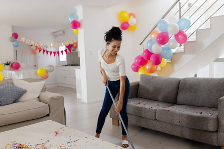 Teenage girl sweeping confetti in modern open-plan living room with balloons, copy space. Cleanup,...