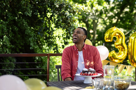 African American man sitting at patio table celebrating thirtieth birthday with cake and balloons. Celebration, festivity, milestone, outdoors, cheerful, elegance, gatheringの写真素材