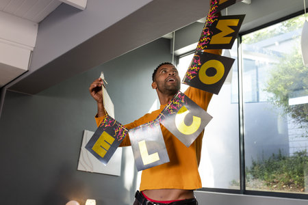 African American man moving WELCOME banner under slanted beam in living room with canvas and lamp....