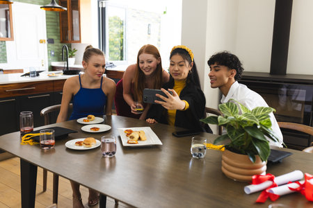 Teens celebrating graduation at home, taking selfie around dining table with snacks. celebration,...