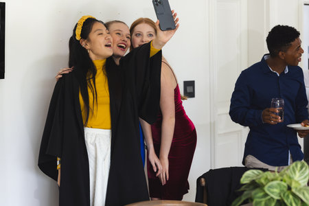Teens celebrating graduation taking selfie at home, smiling and enjoying party. celebration, teenagers, friends, happiness, achievement, diplomaの写真素材