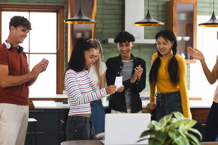 Teen friends celebrating together at home, smiling and clapping around laptop. Friendship, celebration, gathering, technology, happiness, youthの写真素材