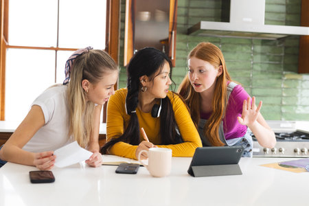 Teen girls collaborating on school project in kitchen, using tablet and notes. Education, teamwork, collaboration, technology, studying, learningの写真素材
